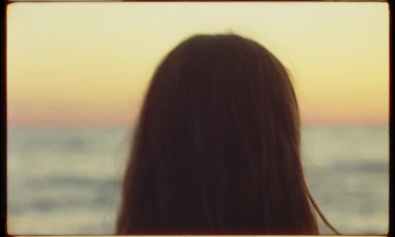 Back view of a young woman with long hair standing by the ocean at sunset slowly turning her head to the side