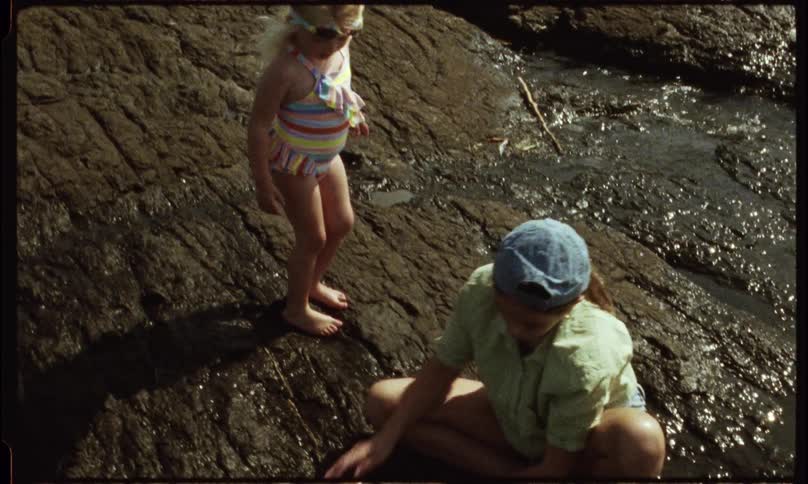 A child in a striped swimsuit stands on wet rocks beside an adult crouching near a shallow stream possibly playing or exploring together outdoors