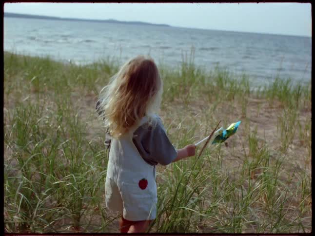 Blonde girl walks towards the sea along the beach filled with grasses holding a paper flower in daytime