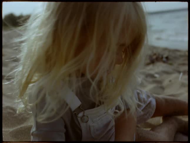 A young child with long blonde hair plays in the sand near a beach uncovering a shiny partially buried object