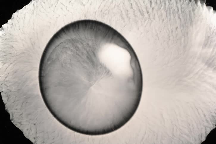 A macro shot shows a dark liquid drop spreading over a textured light surface forming an abstract pattern