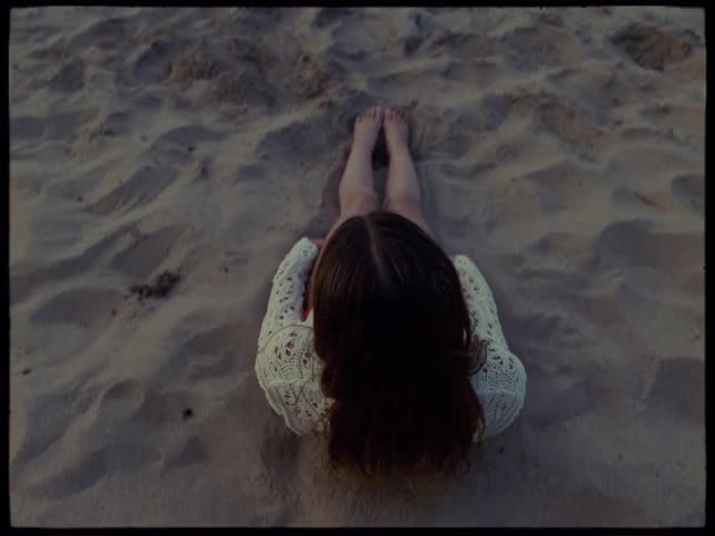 A teenage girl sits up on the beach while waves roll into the shore
