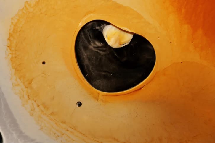 A swirling mixture of black cream and amber liquids forms a hypnotic abstract shape resembling an eye or vortex