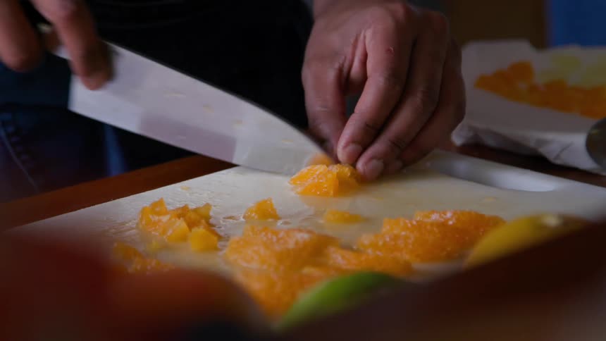The hands of a person using a large knife to cut slices of a yellow food that is on a white cutting board