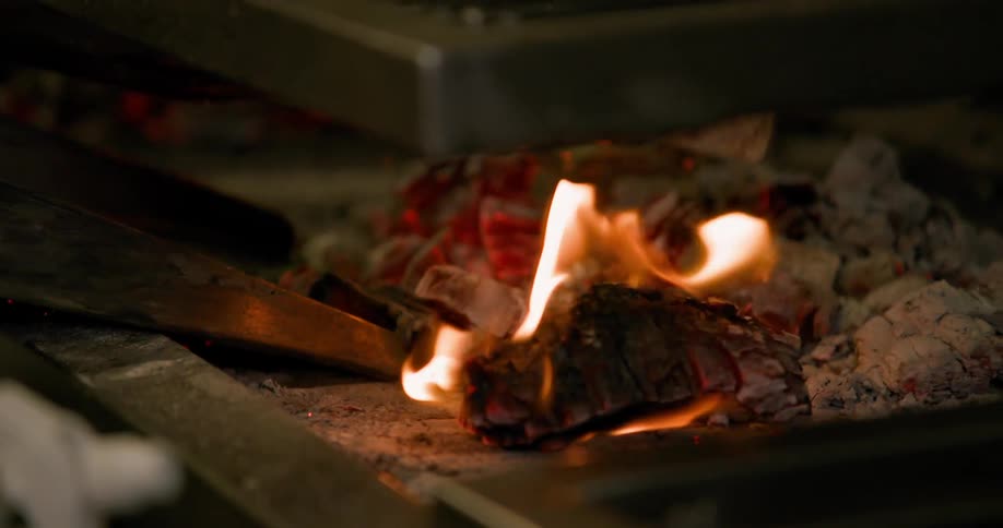 A closeup shot captures the intense flames and glowing embers of a fire burning beneath a grill or metal surface