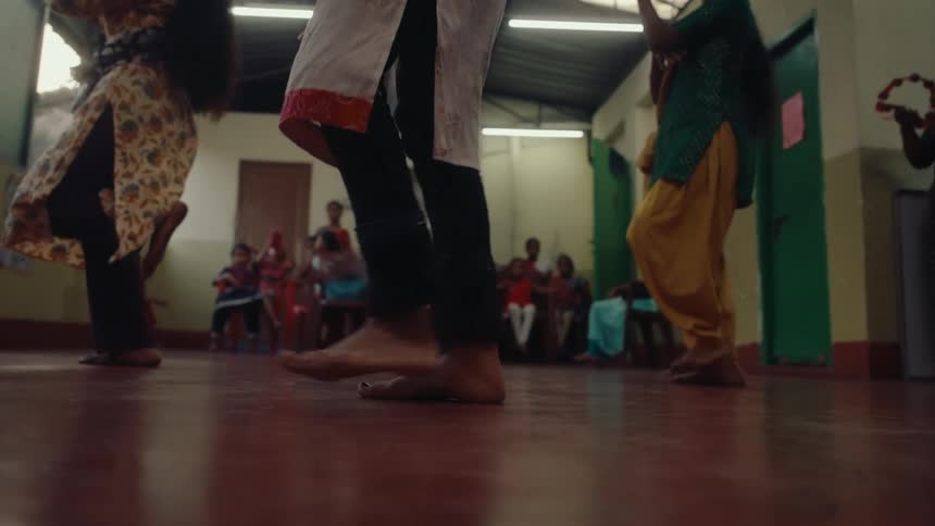 Small group watches barefooted dancers in a building lit by fluorescent lights during the daytime