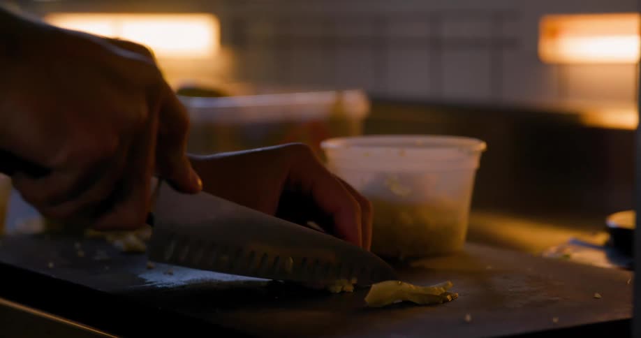 A persons hands are shown using a knife to chop food on a cutting board in a dimly lit kitchen