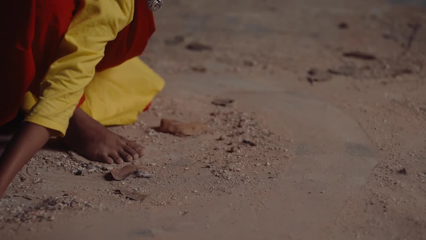 Woman wearing a colorful outfit half kneels while sweeping a hand through the dirt on the ground