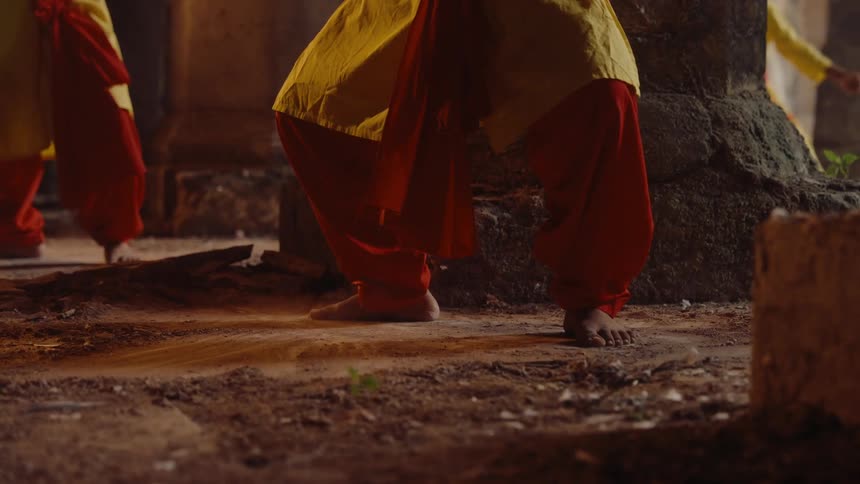 People wearing the same brightly colored outfit stamp bare feet on dust covered ground while standing among stone columns