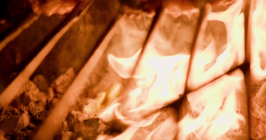A closeup shot shows skewered meat roasting over an open flame with intense heat and fire dominating the frame