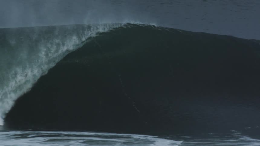 Big wave curls and splashes as it rolls into shore under a dark cloudy sky