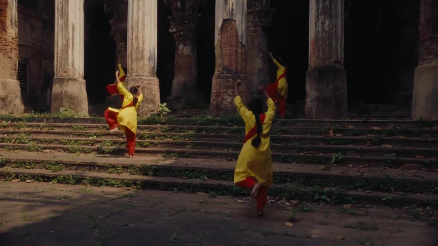 A group of women dressed in coordinated yellow and red traditional outfits energetically dance on the steps of an old weathered stone structure
