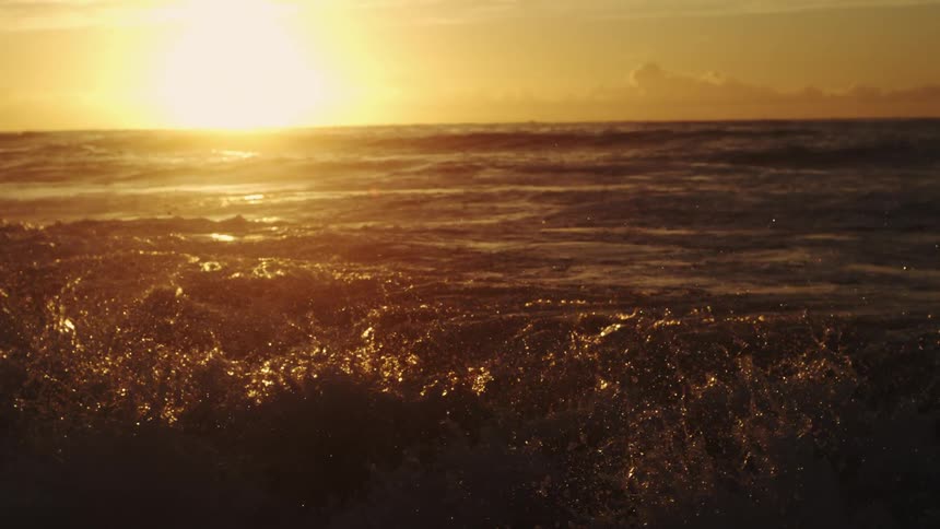 Water splashes as waves roll into shore as the sun shines brightly through clouds on the horizon