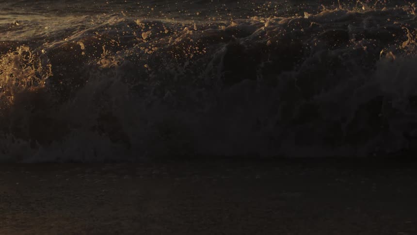 Wave crashes onto the beach while a boat floats near the horizon against the red sky
