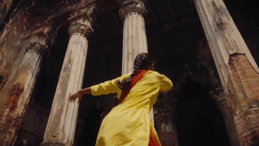Women wearing matching outfits dance near the portico of a large historic building that is going to ruin