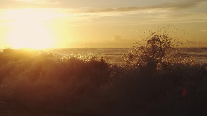 Water splashes as waves roll into shore as the sun shines brightly through clouds on the horizon