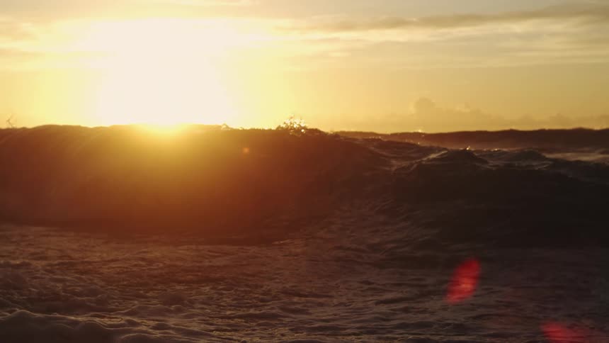 Water splashes as waves roll into shore as the sun shines brightly through clouds on the horizon