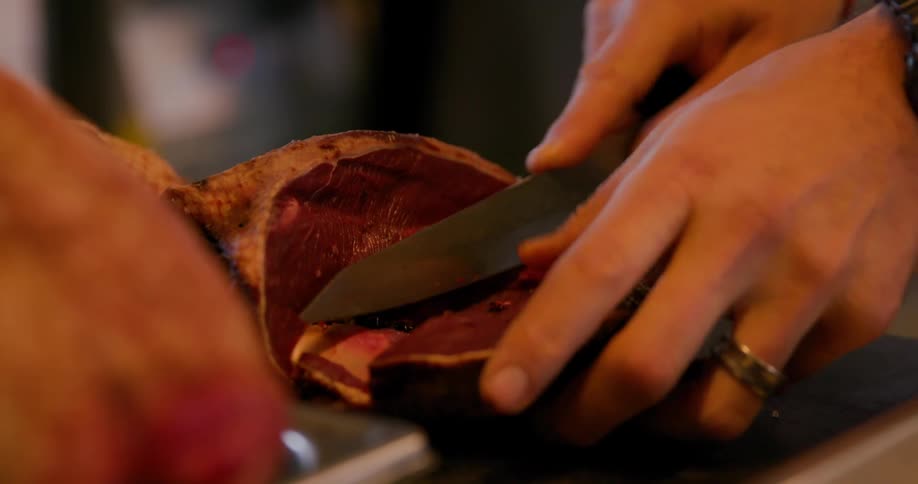 A person is slicing a piece of raw poultry or game meat with a knife shown in a tight closeup kitchen setting