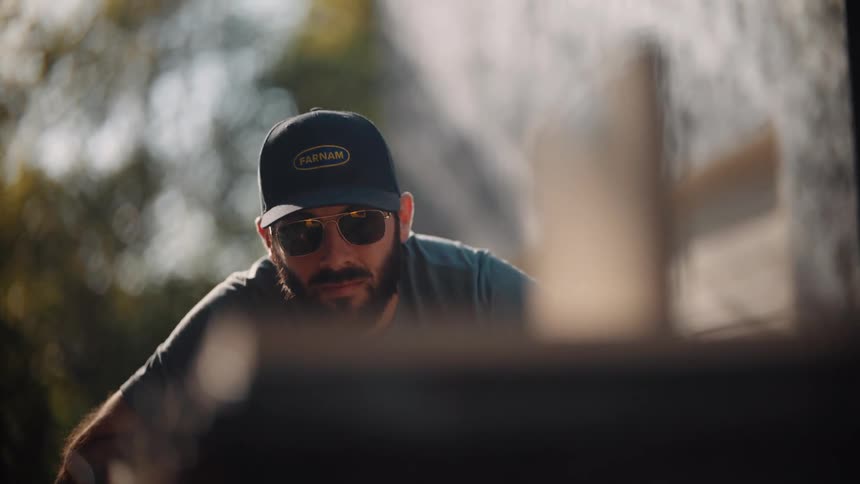 A man in sunglasses and a cap labeled farnam leans forward outdoors in warm sunlight focused and engaged in an activity partially obscured in the foreground