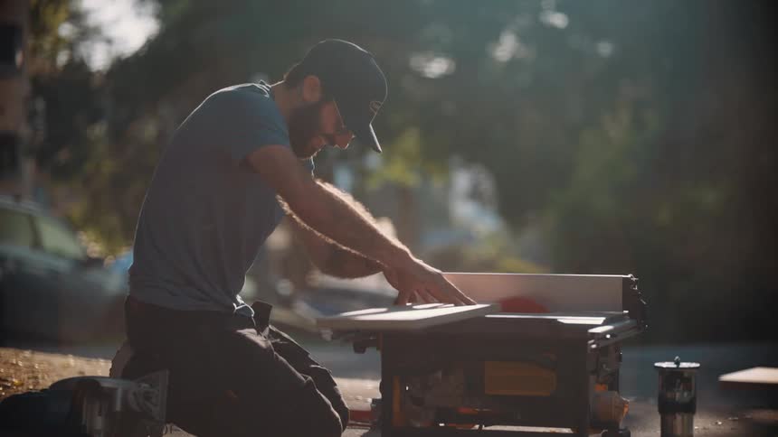 A man kneels outdoors using a table saw to cut a piece of wood