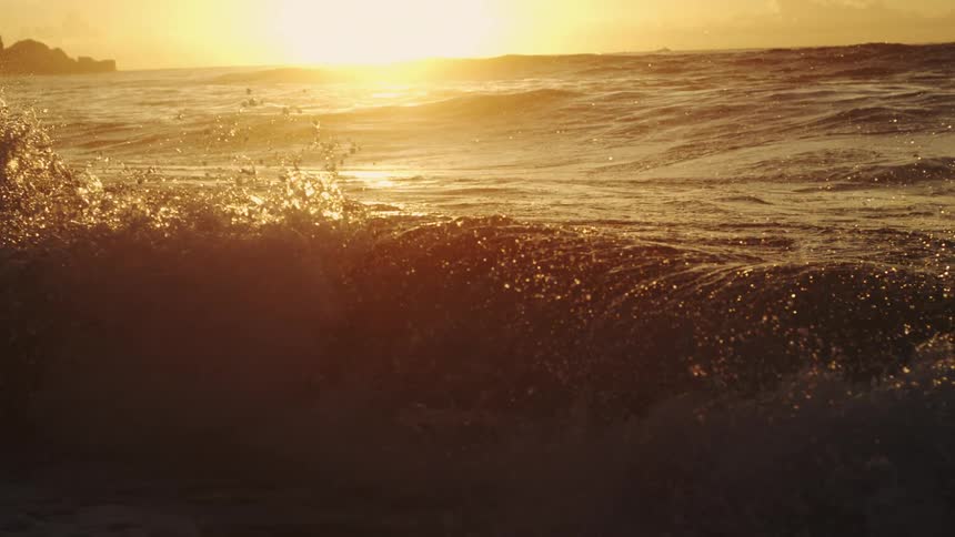 Water splashes as waves roll into shore as the sun shines brightly through clouds on the horizon