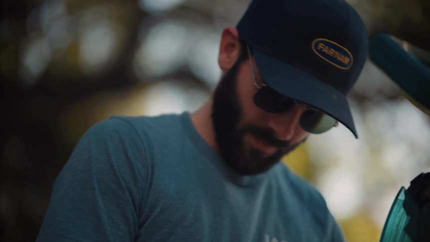 A bearded man wearing a hat with farnam written on the front and sunglasses standing in a park at midday