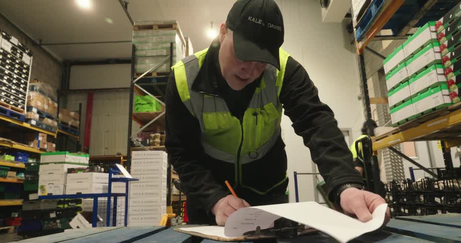 A warehouse worker fills out paperwork while another works in the background among stacked boxes