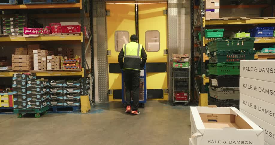 A warehouse worker in safety gear transports boxes of fresh produce using a cart inside an organized storage facility