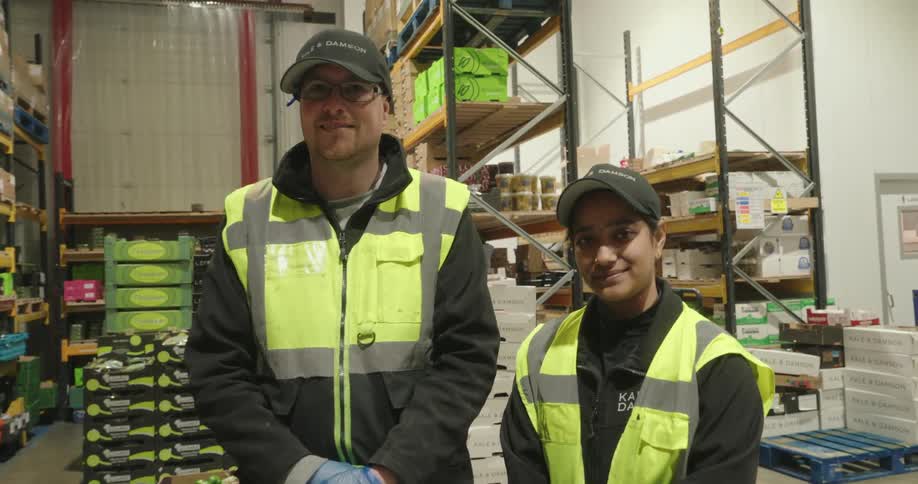 Two workers in highvisibility vests pose and smile inside a warehouse filled with stacked produce and supplies