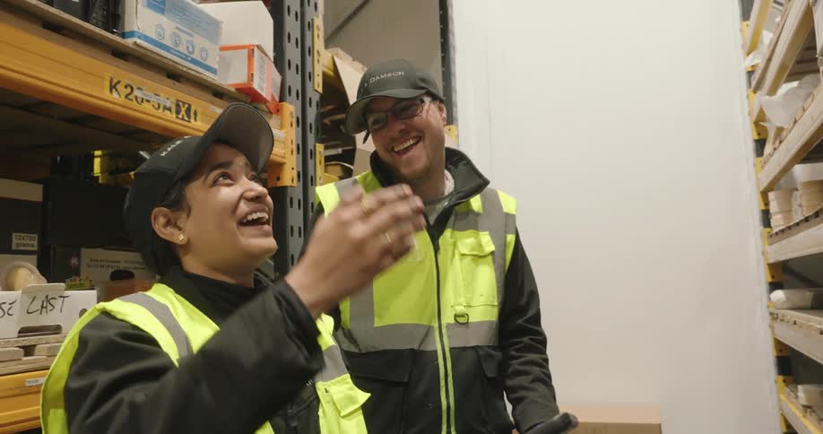 Two warehouse workers in reflective vests smile while looking at a tablet in a storage aisle