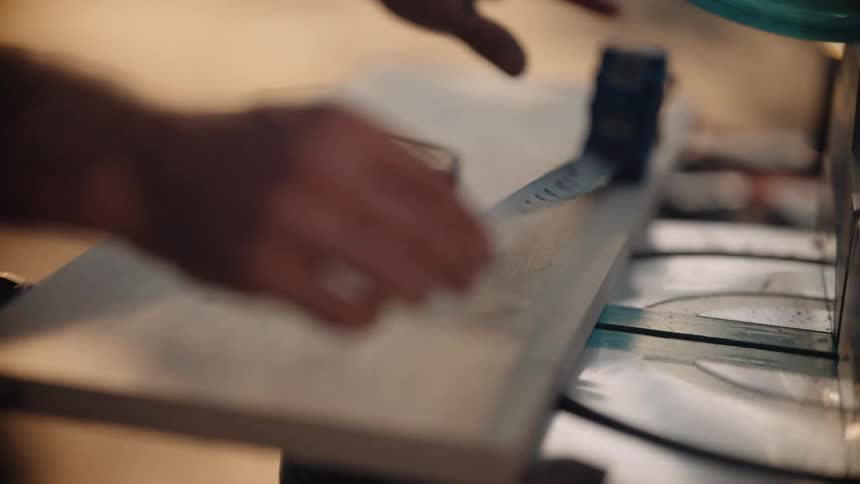Male carpenter measures a piece of wood lying under a table saw
