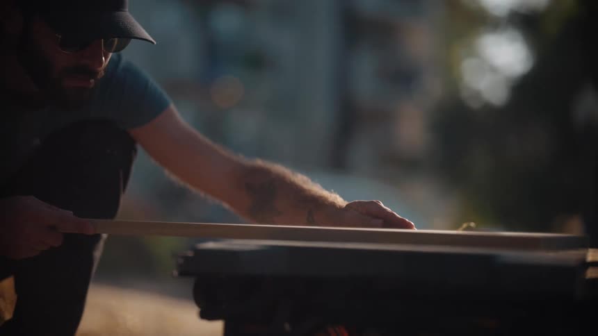 A man wearing sunglasses and a cap is carefully guiding a piece of wood through a machine outdoors in soft moody lighting
