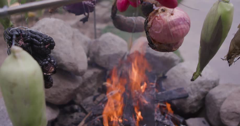 Various vegetables including corn onion and peppers are suspended over an open flame for roasting in an outdoor setting