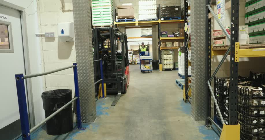 A warehouse worker in a highvisibility vest walks near a forklift amid shelves stacked with boxes