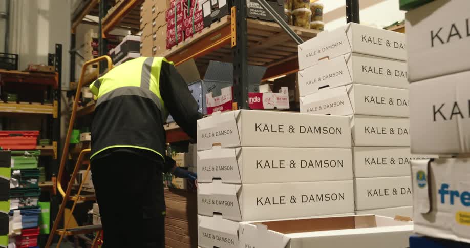 A worker in a highvisibility vest is handling and organizing produce boxes inside a commercial storage or warehouse facility