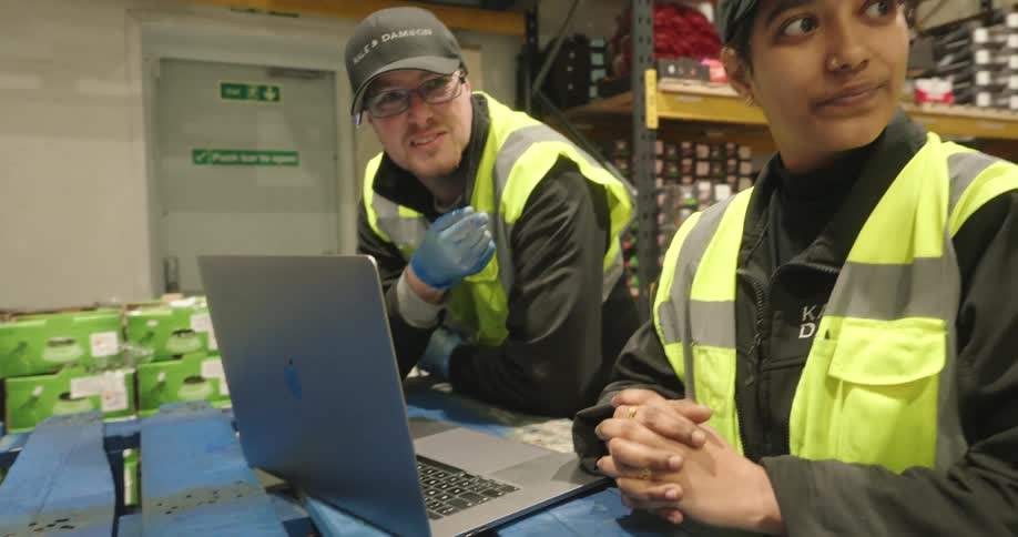 Two warehouse workers in highvisibility vests smile and collaborate while looking at a laptop on a pallet inside a storage facility