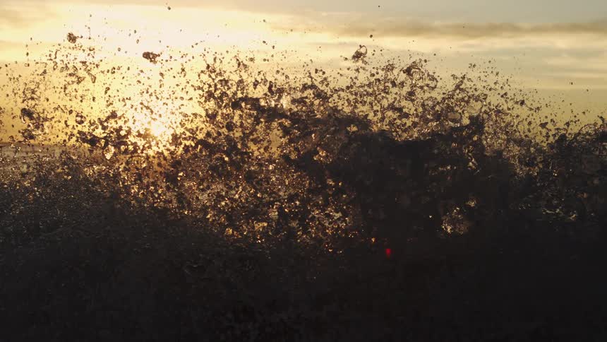Spray from a wave crashing onto the beach partially obscures the sun shining on the horizon