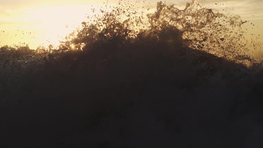 A slow motion shot of an ocean wave crashing back into the water with the sun rising far on the horizon and a hazy cloudy sky hovering over the turbulent waters