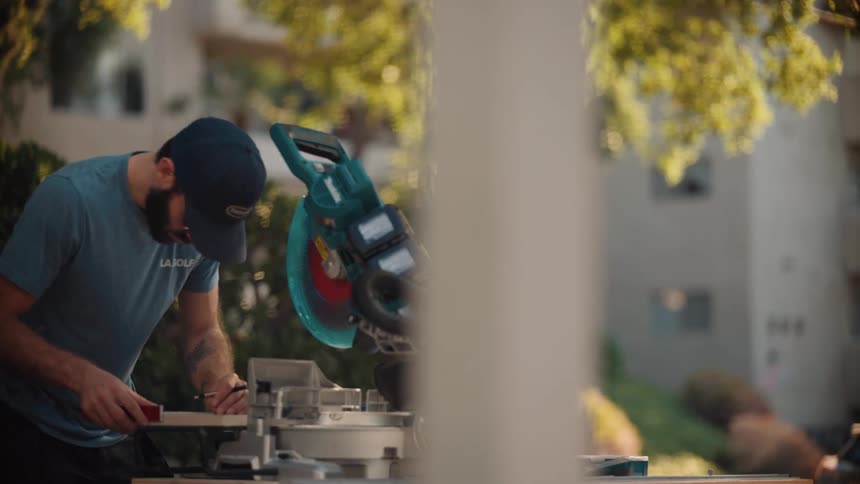 A man in a blue shirt and cap is focused on using a compound miter saw outdoors in a sunlit residential area
