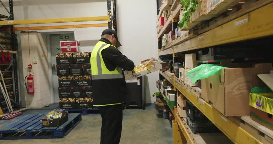 A male worker inside a warehouse is sorting items on a shelf wearing a green safety vest and a hat
