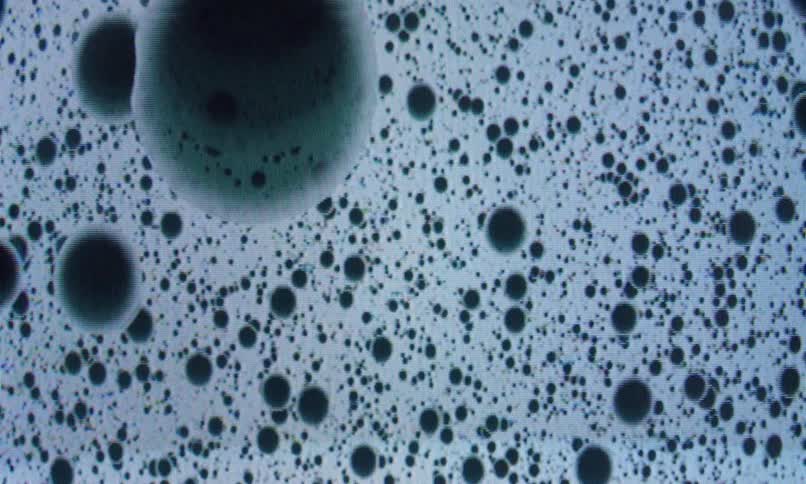 A highly magnified abstract visual of dark circular shapes resembling droplets or particles suspended in a light blue fluid background