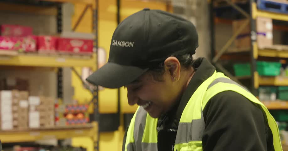 Three workerstwo male and one femalewearing safety vests and hats are talking and laughing together inside a warehouse