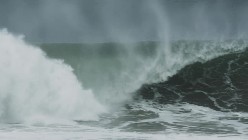 Surfers surf an ocean wave that approaches and crashes against the shore in day time