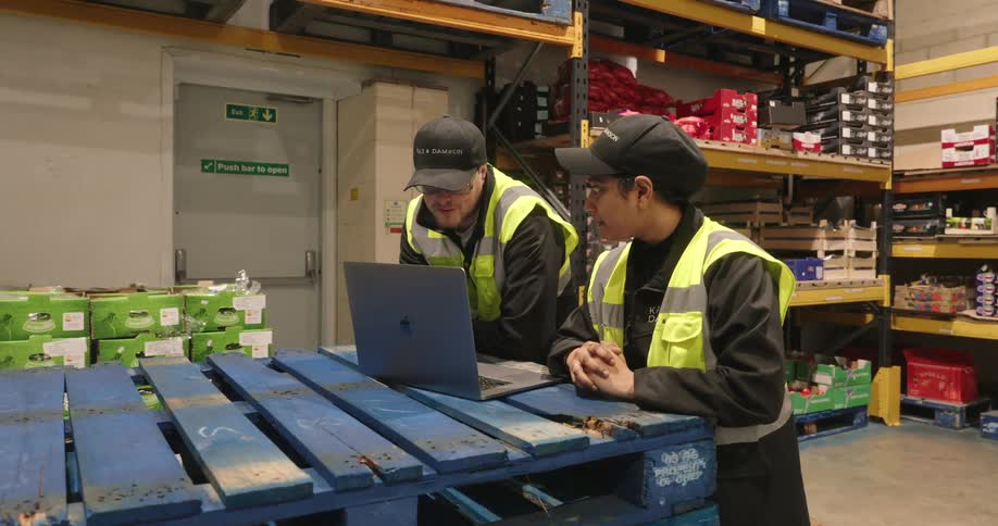 Three workers in high visibility vests are using a laptop on a pallet in a warehouse filled with shelves of goods