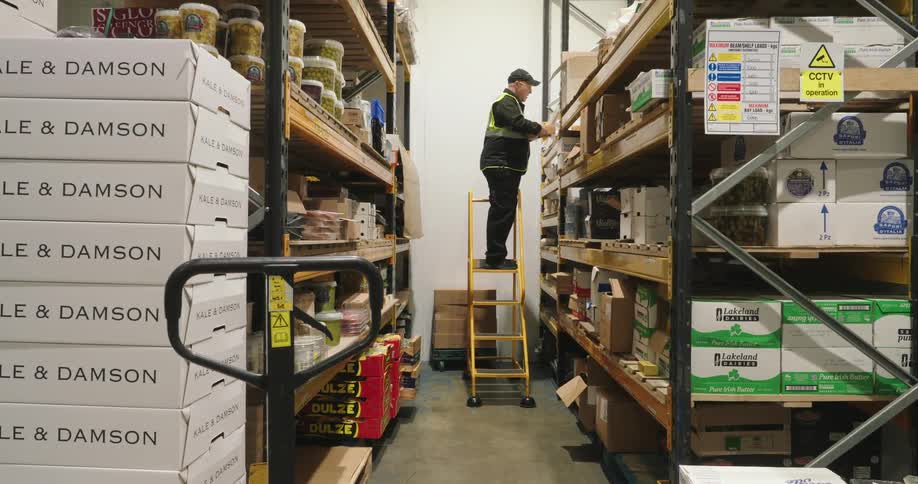 An employee in a warehouse climbs up a platform ladder to put tape on a box