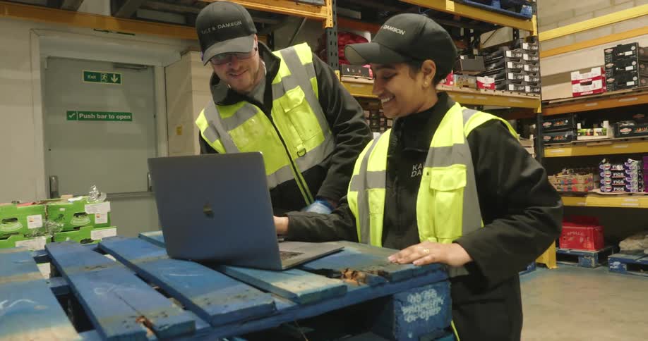 One employee shows another employee something on a laptop inside of a warehouse