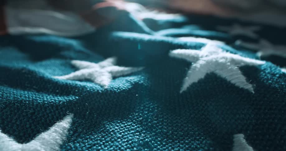 A closeup shot captures the textured fabric of an american flag with a lens flare illuminating the upper right corner