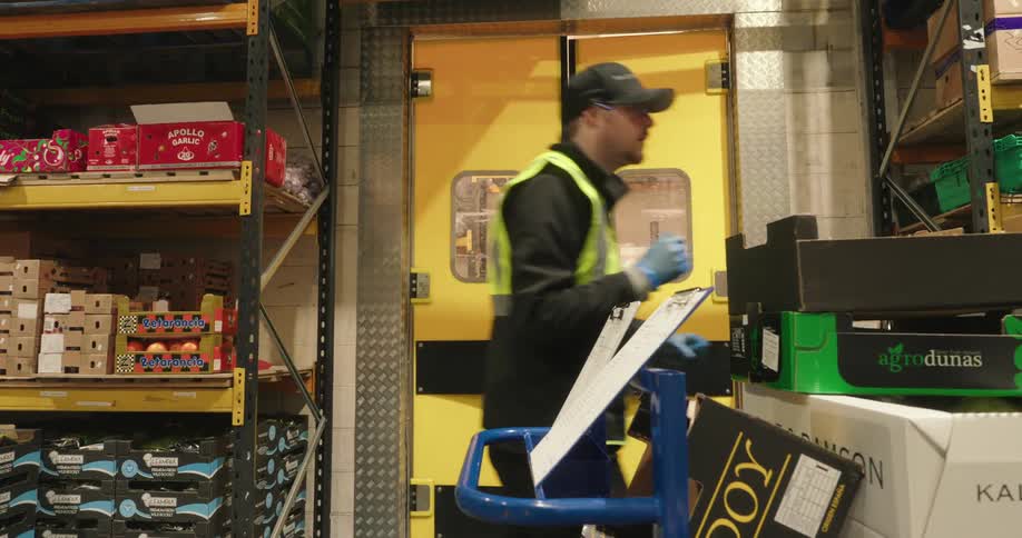 The worker checks his clipboard and goes over to the shelf