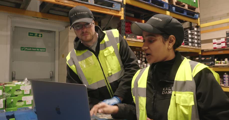 A woman and a man talking in a warehouse with the woman explaining something she sees on her laptop