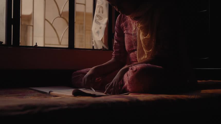 A woman sits beside a window at midday holding a pen as she scans through a notebook in her hands